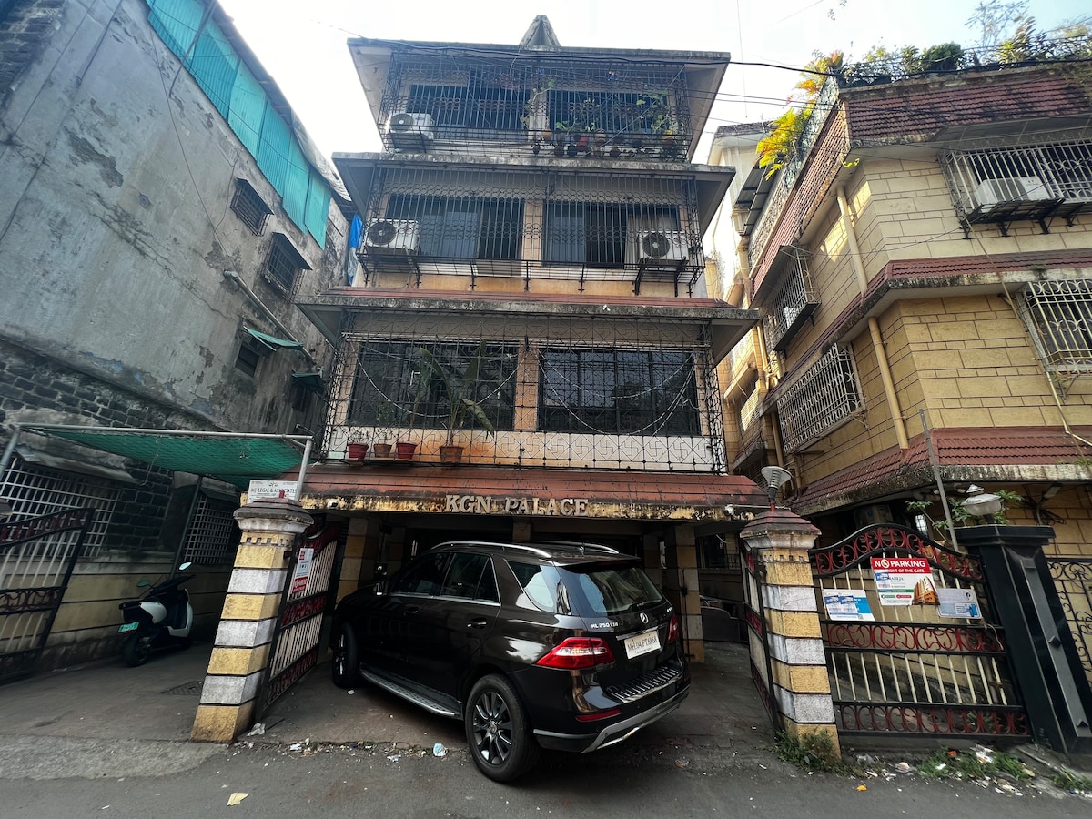 A multi-story building is featured, showcasing a facade with decorative railings and multiple air conditioning units installed on the exterior. A parked SUV is positioned beneath a shaded overhang, creating an inviting entryway. The surrounding area includes glimpses of neighboring structures.