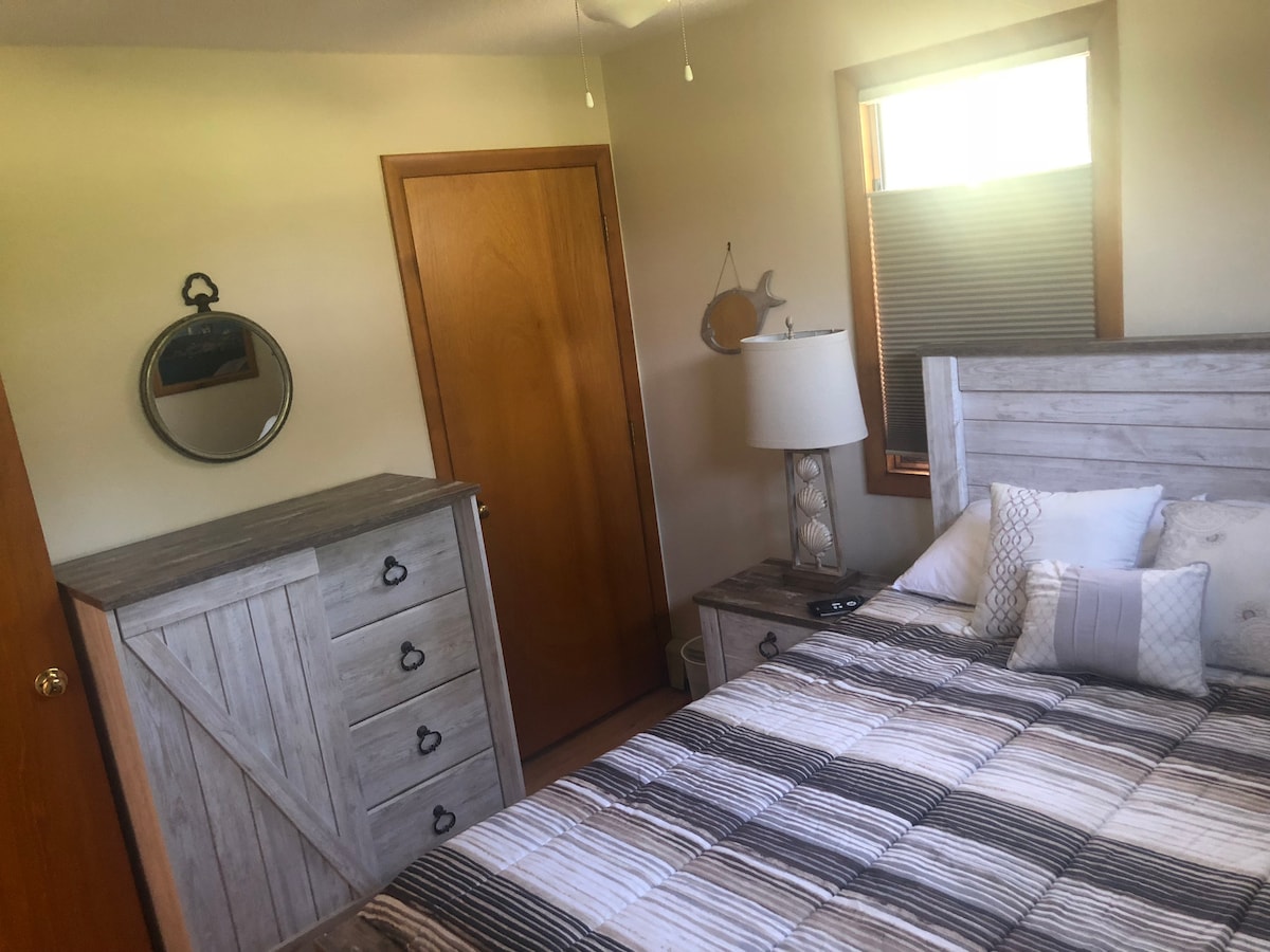 A cozy bedroom features light-colored wooden furniture and a spacious bed with a striped comforter. A round mirror is mounted on the wall above a dresser. Natural light filters through a window, and a door leads to the hallway.
