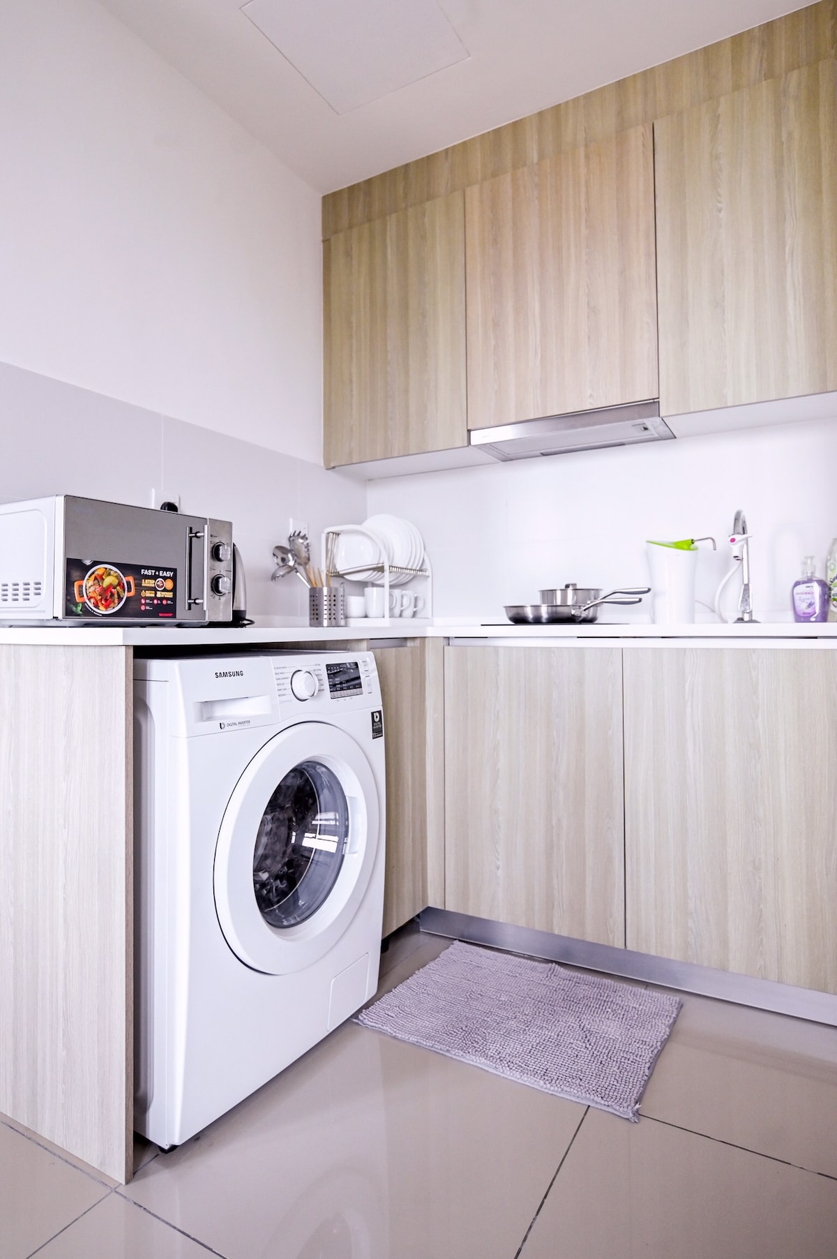 A modern kitchen area features light wooden cabinetry and a compact countertop. Equipped with a washing machine, microwave, and various cooking utensils, the space is designed for convenience. A soft grey mat is positioned in front of the washing machine on the polished floor.