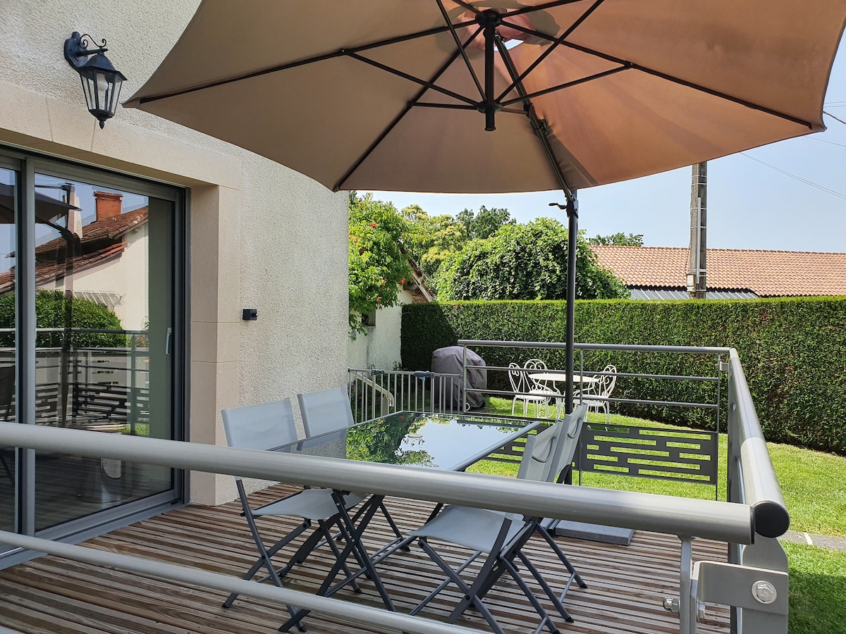 A terrace features a large glass-top table surrounded by several chairs, shaded by a large umbrella. A barbecue grill is positioned nearby, and a privacy hedge creates a serene outdoor space that invites relaxation.