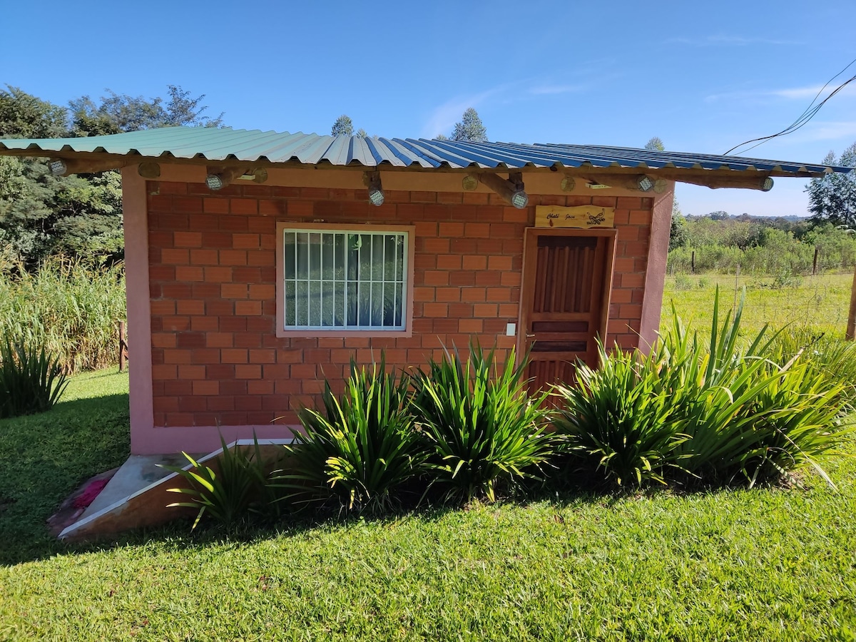 A rustic chalet features a brick exterior with a green metal roof. A wooden door is centered under a decorative sign, while a large window allows light to enter. Lush green grass and ornamental plants surround the structure, enhancing its natural setting.