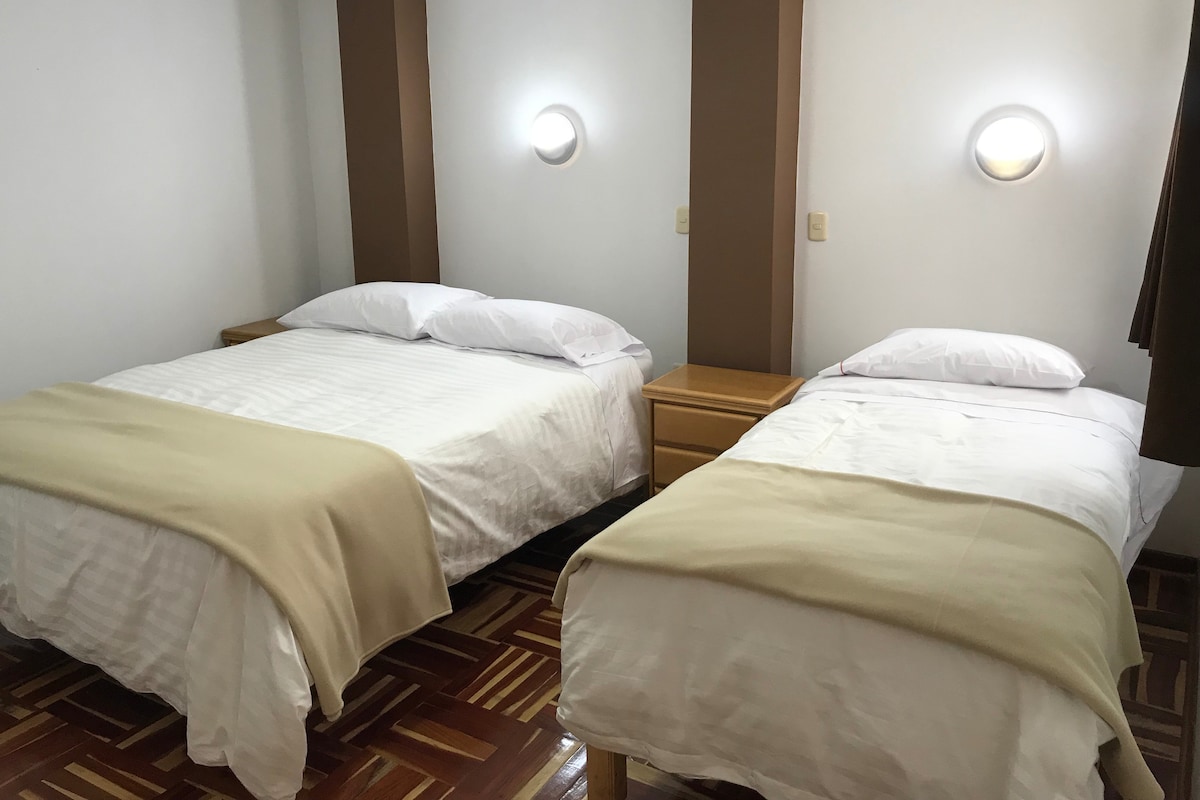 Two neatly arranged beds are positioned in a well-lit room, one bed featuring a double size and the other a single. Soft beige blankets complement the crisp white linens. Wooden nightstands flank each bed, enhancing the functional and warm atmosphere.