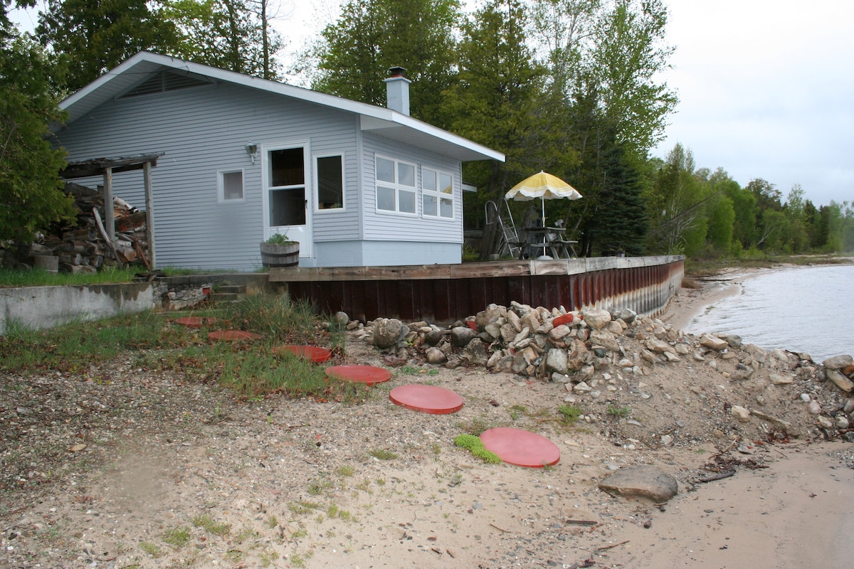 A serene lakeside retreat is shown, featuring a light blue exterior and large windows. A wooden deck extends towards the water, furnished with a small table and chairs under an umbrella. Smooth stones and a sandy shoreline create a natural path to the water’s edge.