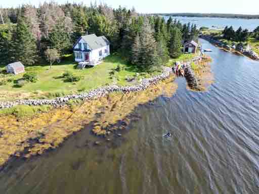 An aerial view captures the private island cottage surrounded by lush greenery and water. The blue and white character house is visible along with a stone path leading to the private wharf. Calm waters reflect the shoreline, offering a serene natural environment.