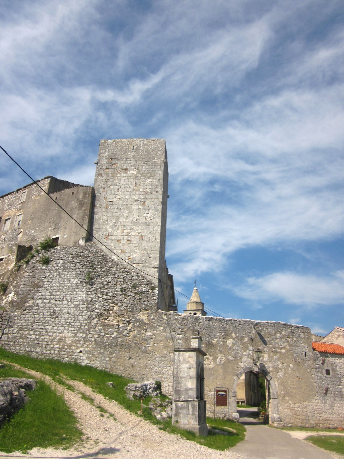 A historic stone structure rises against a blue sky, featuring a tall tower and a partially visible gate. The surrounding area is lush with grass, and a pathway leads towards the entrance, inviting exploration of the fortifications and landscape.