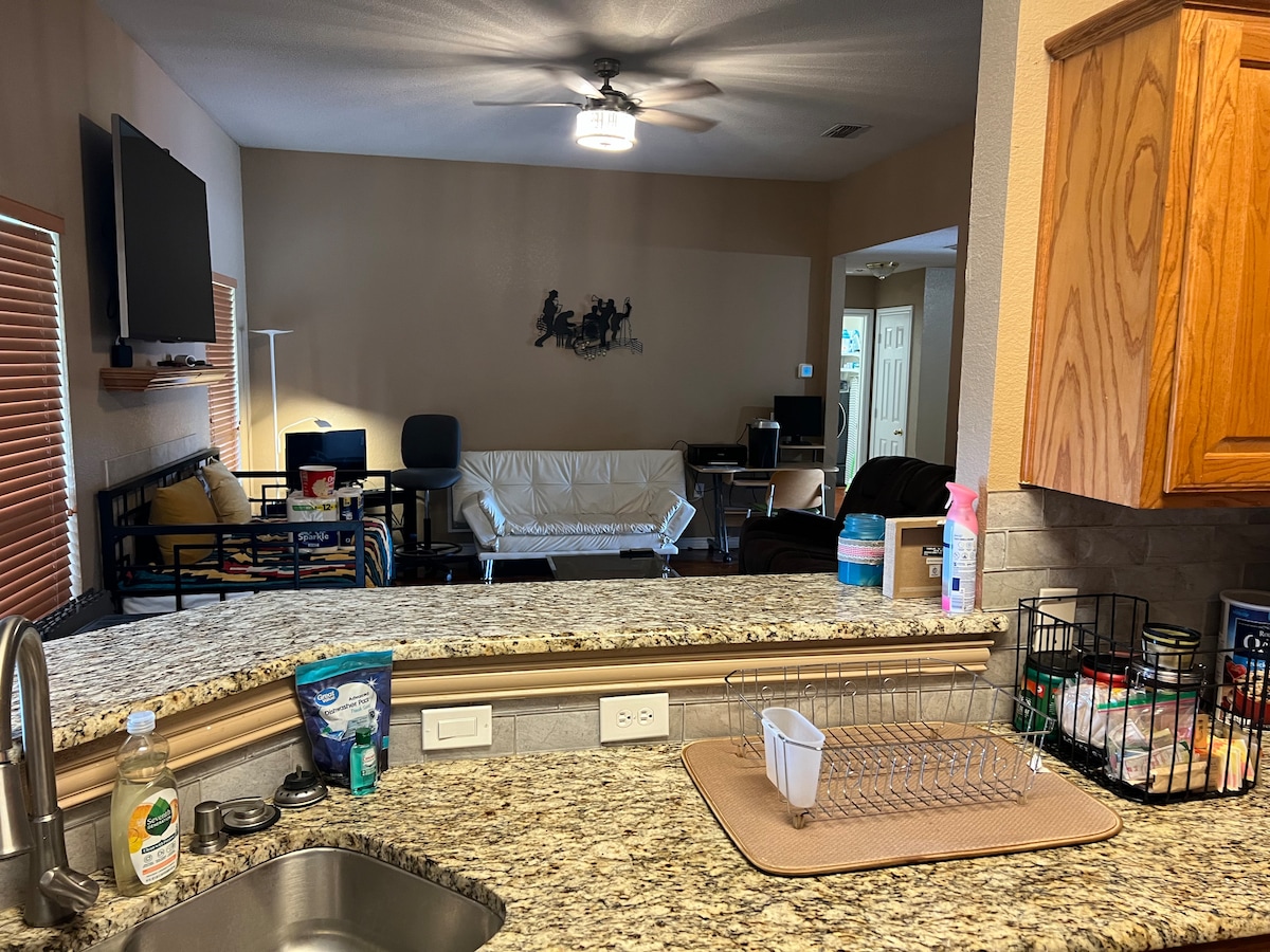 The kitchen is equipped with granite countertops and a stainless steel sink, offering a view of a spacious living area. A mounted television is visible, along with seating arrangements that include a white futon and a dark-colored couch. Cabinets hold various pantry items and cleaning supplies.