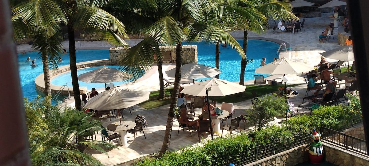 A scenic pool area is surrounded by lush palm trees, featuring several loungers and umbrellas. Multiple swimming pools in varying sizes can be seen, with guests enjoying the water. A well-maintained garden enhances the atmosphere, creating a relaxing outdoor space.
