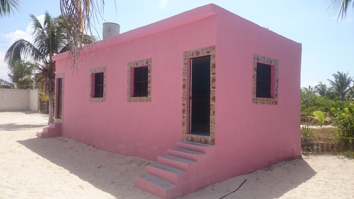 A pink house with a simple design is presented, featuring three windows with iron bars and two steps leading to the entrance. The sandy surroundings suggest proximity to the beach, complemented by palm trees in the background, enhancing the appeal of the tranquil setting.