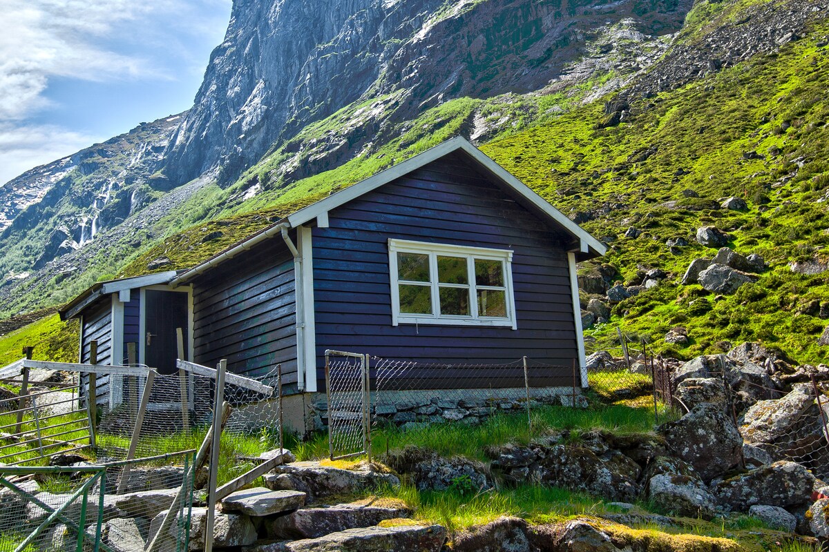 A mountain cabin is nestled against a rocky hillside, surrounded by lush green grass and boulders. The wooden structure features large windows and a sloped roof. A simple fence delineates the area, complementing the natural landscape of steep mountains and clear skies.