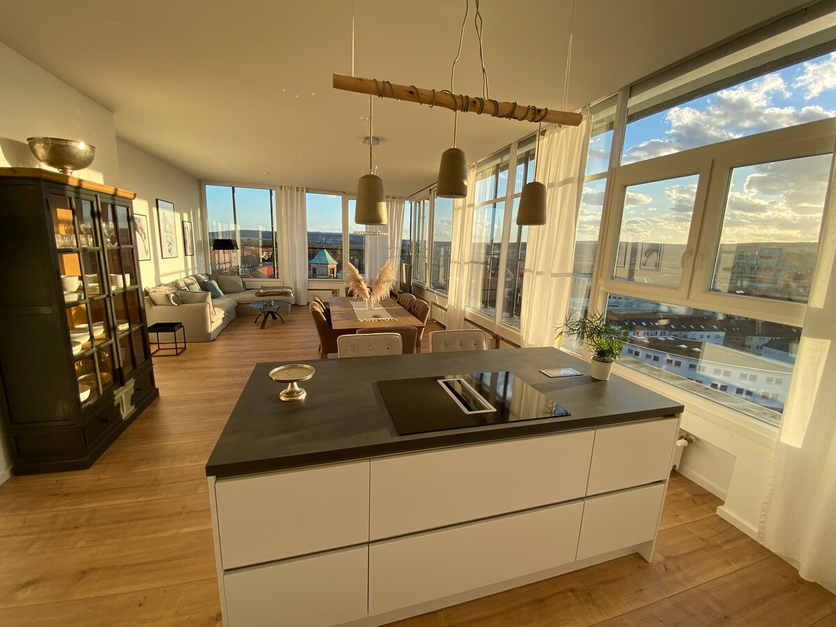 A modern kitchen features a sleek island with a cooktop and minimalist cabinetry. Large windows allow natural light to illuminate the adjoining living area, showcasing comfortable seating and scenic views. The open layout enhances connectivity throughout the space.
