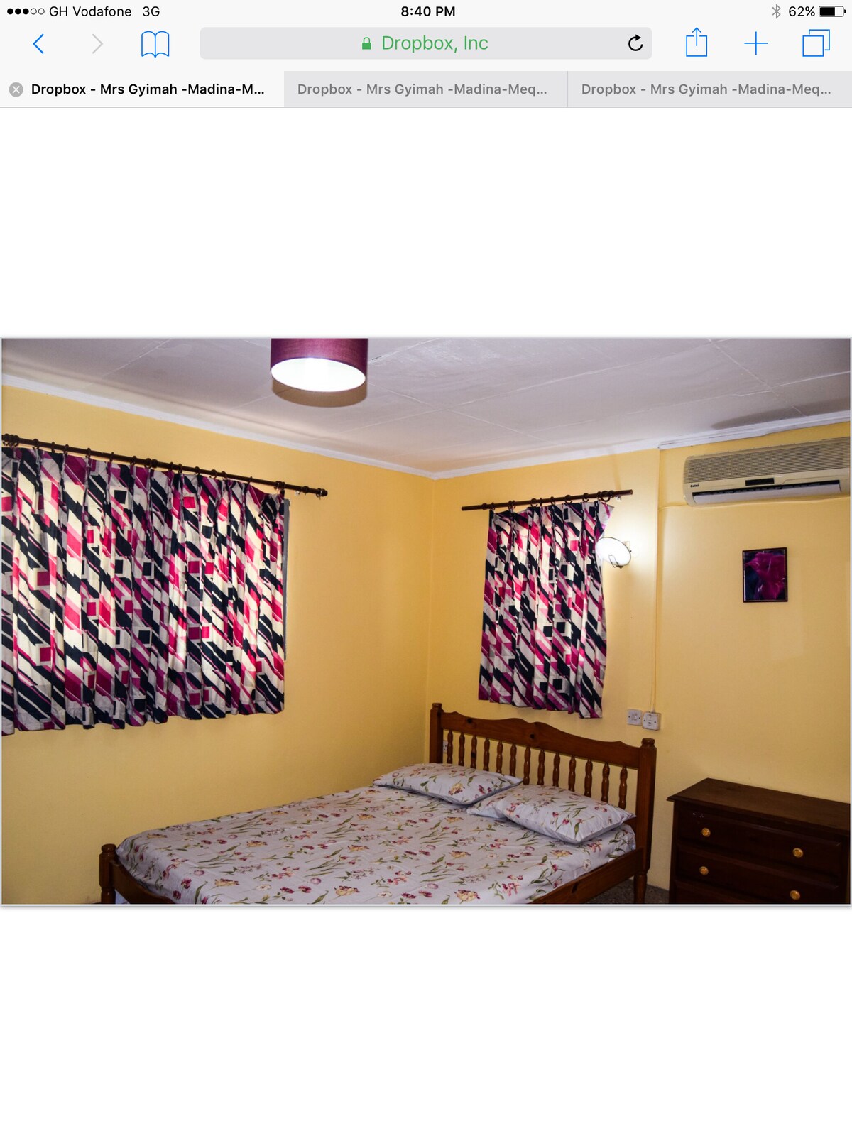 A cozy bedroom features a wooden bed with a floral patterned duvet. Two pillows are placed at the head of the bed. Bright yellow walls are complemented by patterned curtains, while an air conditioning unit is mounted above the bed, adding comfort to the space.