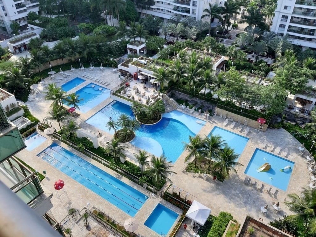 An aerial view showcases a vibrant complex featuring multiple swimming pools surrounded by lush greenery. The layout includes various shapes of pools, along with shaded seating areas and palm trees, creating a relaxing outdoor environment. Lounge chairs are positioned around the pools for leisure.