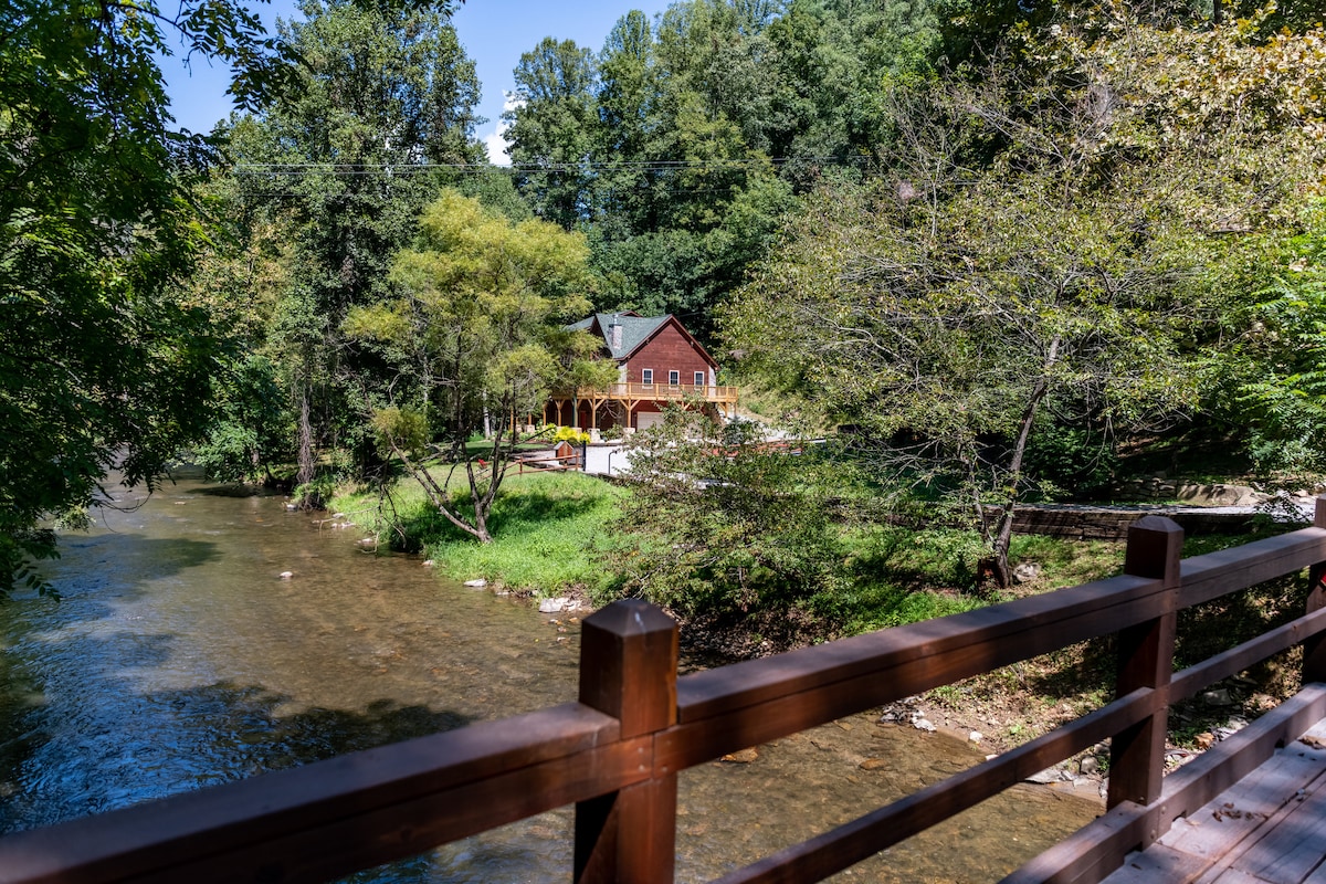 The cabin is nestled alongside a tranquil creek, with lush greenery surrounding it. A spacious deck can be seen, providing a scenic view of the water and landscape. Afternoon sunlight filters through the trees, creating a calming atmosphere by the water's edge.