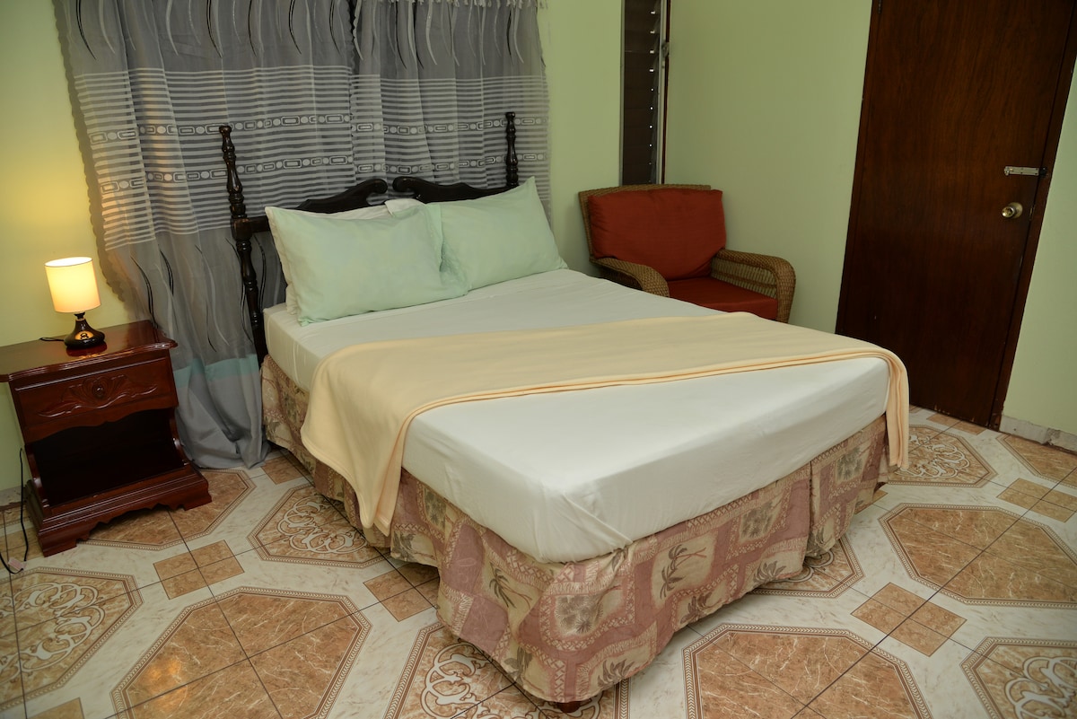A cozy room is equipped with a full bed adorned with light-colored pillows and a soft blanket. A wicker chair is positioned near the bed, and warm light from a bedside lamp illuminates the space. Decorative curtains frame a window, adding to the serene atmosphere.