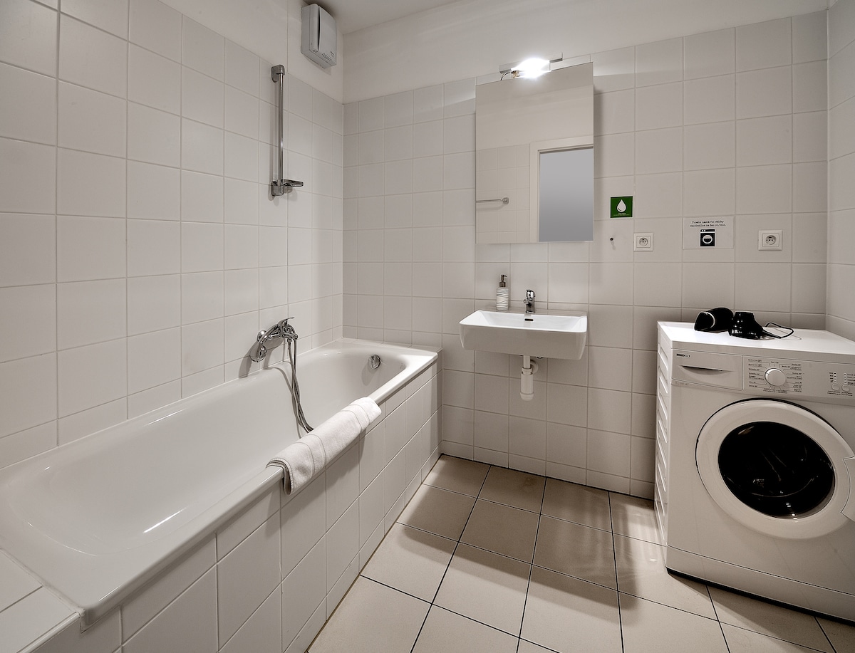 A clean, well-lit bathroom features a bathtub and a modern sink mounted on a white wall. A mirror is positioned above the sink. A washing machine is present, providing convenience for guests. Light-colored tiles cover the floor, enhancing the spacious feel.