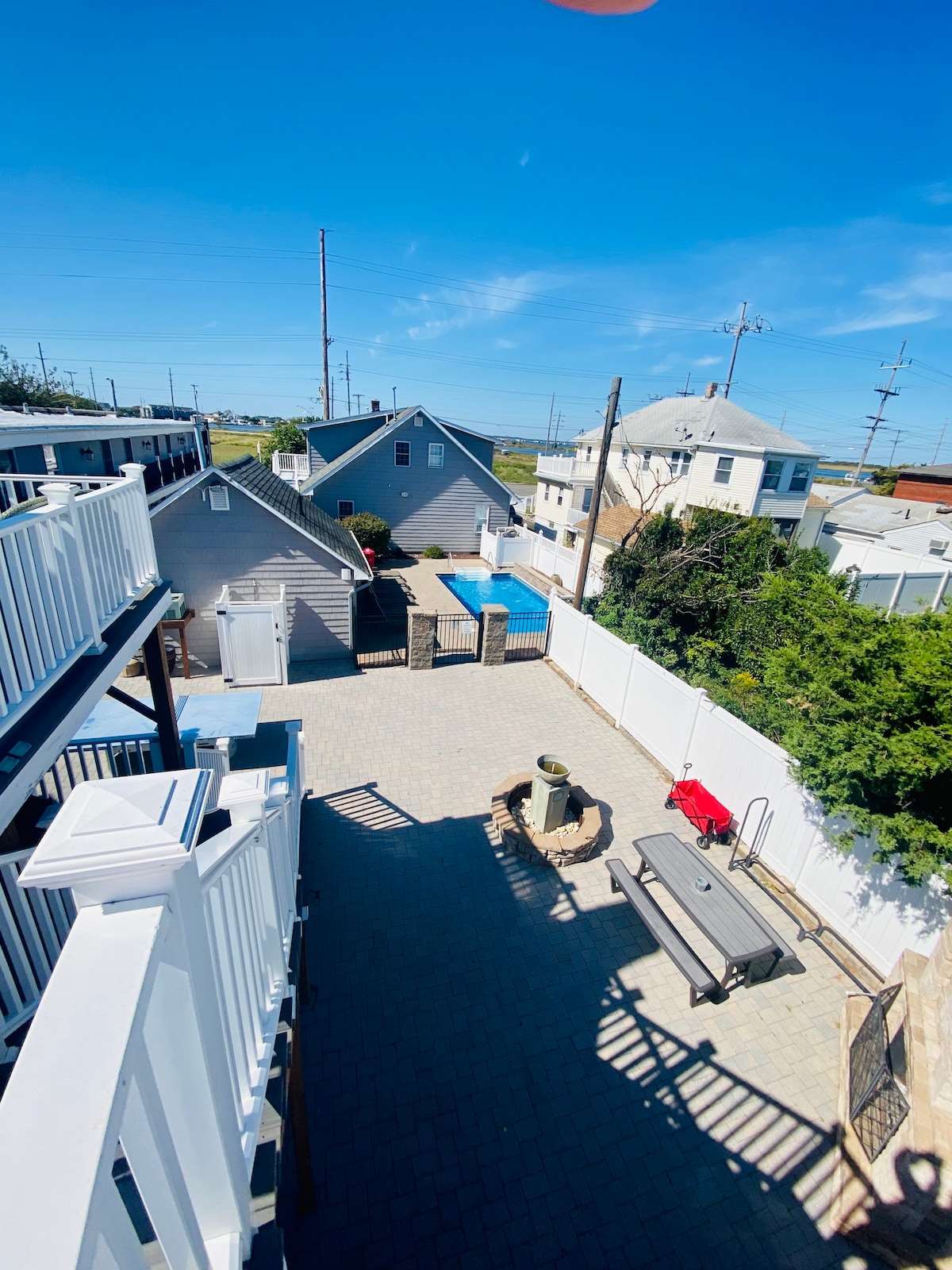 A spacious outdoor area is shown, featuring a paved surface and distinct boundaries marked by white fencing. One in-ground pool is visible along with various nearby structures. A BBQ area and outdoor furniture are set up, creating an inviting environment for relaxation and gatherings.