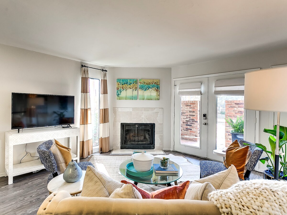 A spacious living area features a light-colored sofa adorned with decorative pillows, positioned around a glass coffee table. A flat-screen TV is mounted on a white console. Large windows allow natural light to fill the room, complemented by sliding doors leading to an outdoor space.
