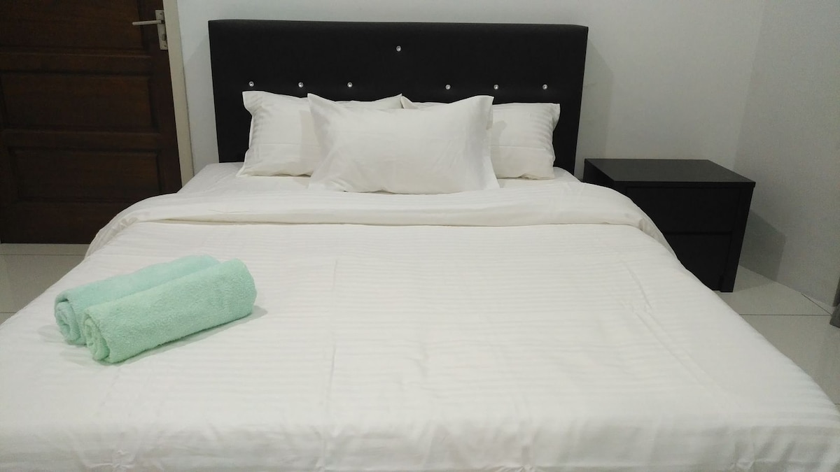 A spacious bed is featured prominently in the image, adorned with crisp white linens and several plush pillows. Two rolled towels in a soft aqua color are positioned neatly at the foot of the bed. A bedside table is visible to the side.