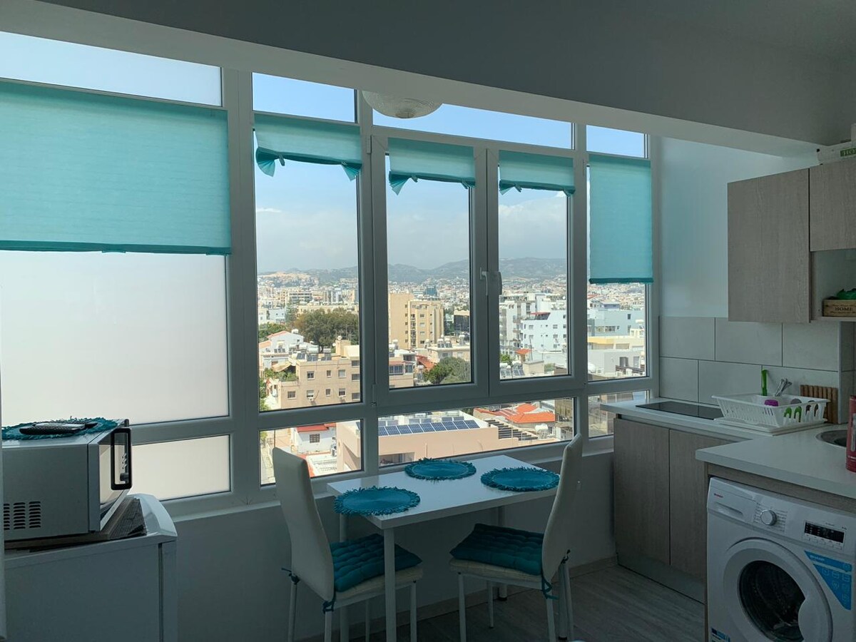A bright kitchenette is bathed in natural light from large windows with sheer turquoise shades. A small dining table with two chairs is positioned adjacent to a washing machine and kitchen counter. Scenic views of the city and mountains are visible outside.