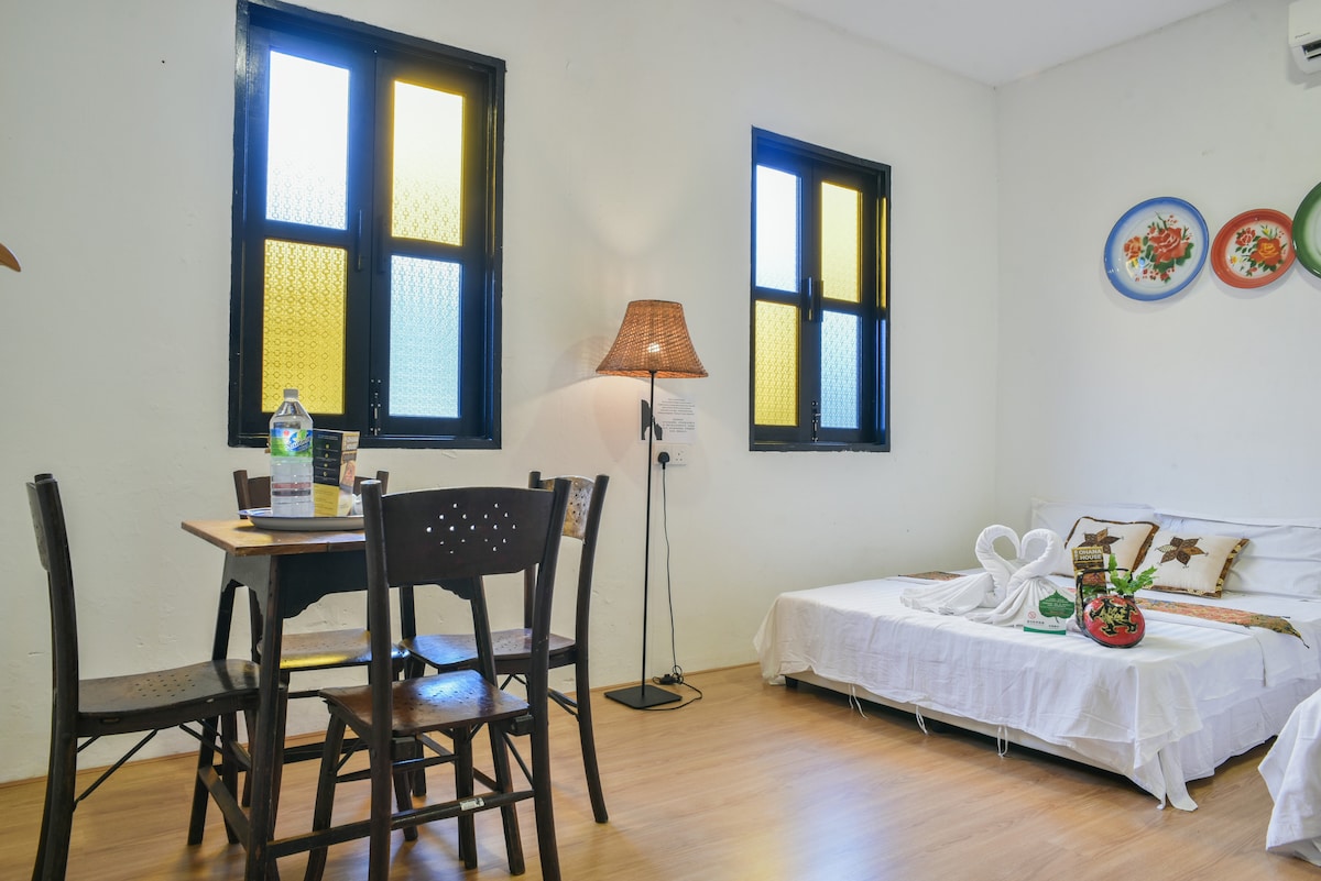 A bright room features two windows with yellow and clear glass. A small dining table with four chairs sits beside a neatly made bed. A lamp provides soft lighting, while decorative plates adorn the wall, creating a cozy atmosphere.