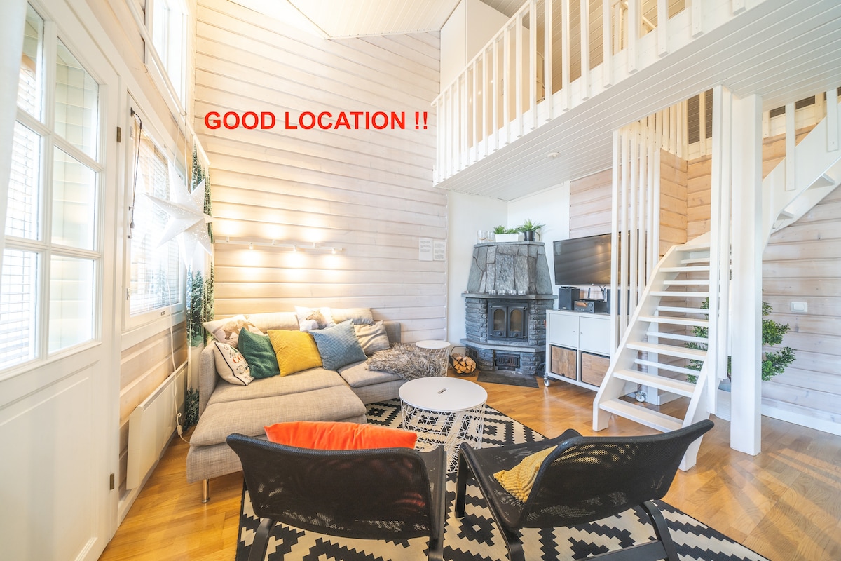 The living area features a spacious seating arrangement with a sectional sofa adorned with colorful cushions. A circular coffee table is positioned in the center, and a television stands beside a cozy fireplace. A staircase leads to an open upper floor, with ample natural light from large windows.