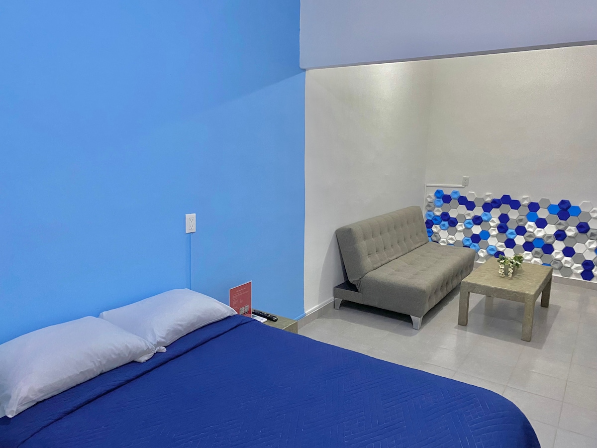 A minimalist room features a comfortable bed with blue bedding and two pillows, alongside a modern gray sofa. A small table sits near the seating area. The walls are painted a calming blue, and the design includes decorative tiles that add a touch of color.