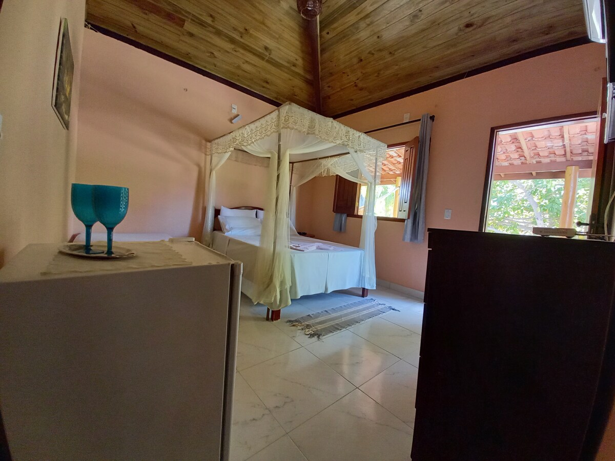 The room features a spacious bed with a decorative canopy, complemented by light-colored bedding. Natural light flows in through the windows, which are adorned with simple curtains. The ceiling showcases wooden beams, enhancing the rustic charm of the accommodations.