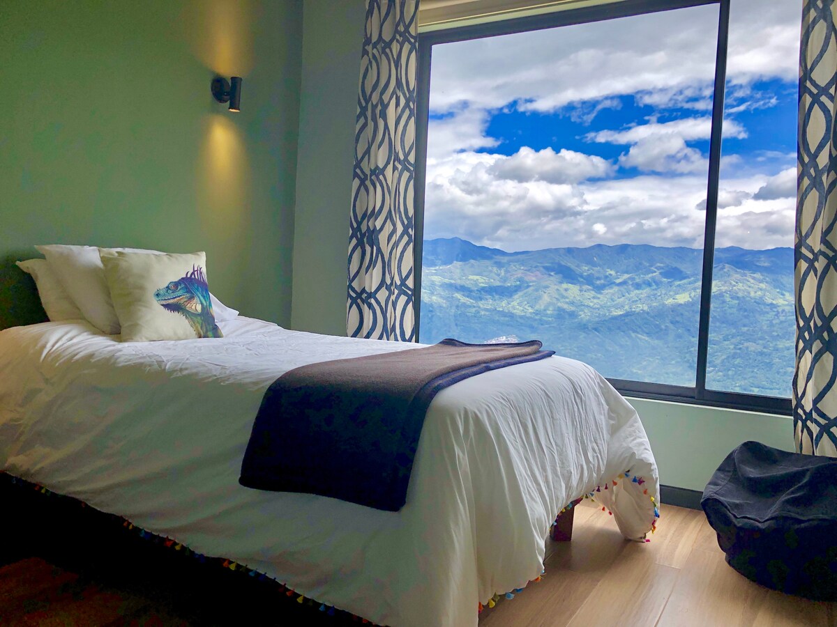 A cozy sleeping area features a spacious bed, complemented by decorative pillows and a soft blanket. Large windows showcase stunning views of the surrounding mountains and sky, while natural light fills the room, highlighting the calming green walls.