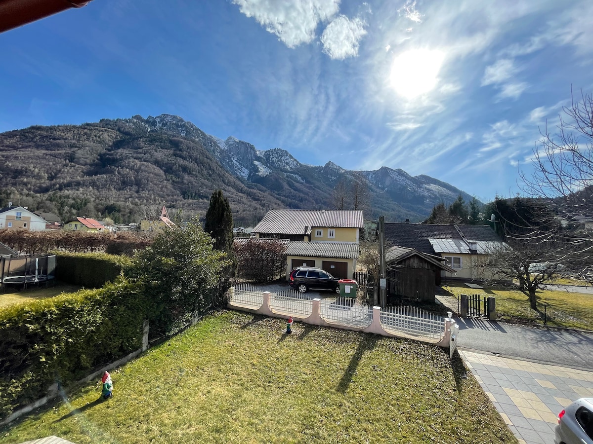 The image showcases a scenic view of the surrounding landscape, featuring towering mountains in the background. A well-kept garden with green grass and decorative elements is visible in the foreground, while a suburban area with houses lies further back, along with a bright sun in a clear sky.