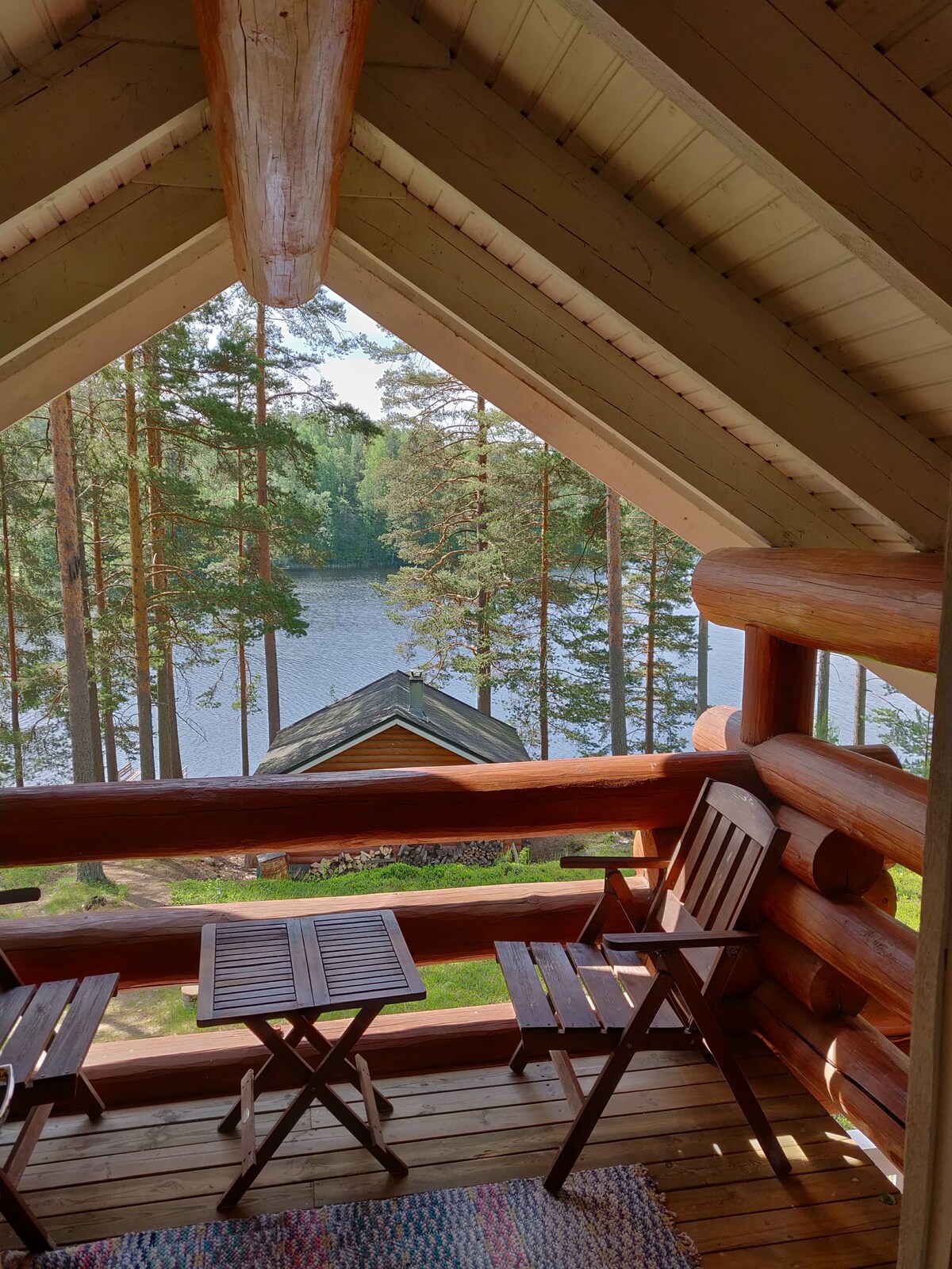 A view is captured from a balcony with wooden railings, featuring two foldable chairs and a small table. Trees surround the tranquil water below, while a rustic building is visible in the distance, framed by the sloped roofline.
