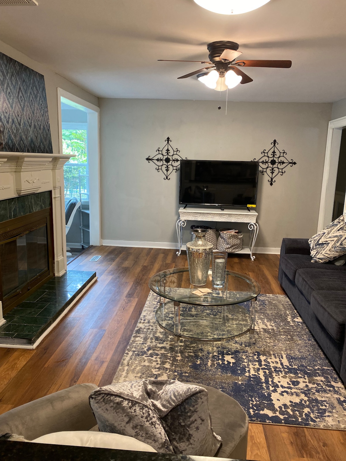 A living room features hardwood flooring and a dark sofa facing a mounted television. A decorative glass coffee table sits on a patterned rug, complemented by metallic vases. The fireplace is styled with green marble, and natural light filters in from a nearby window.