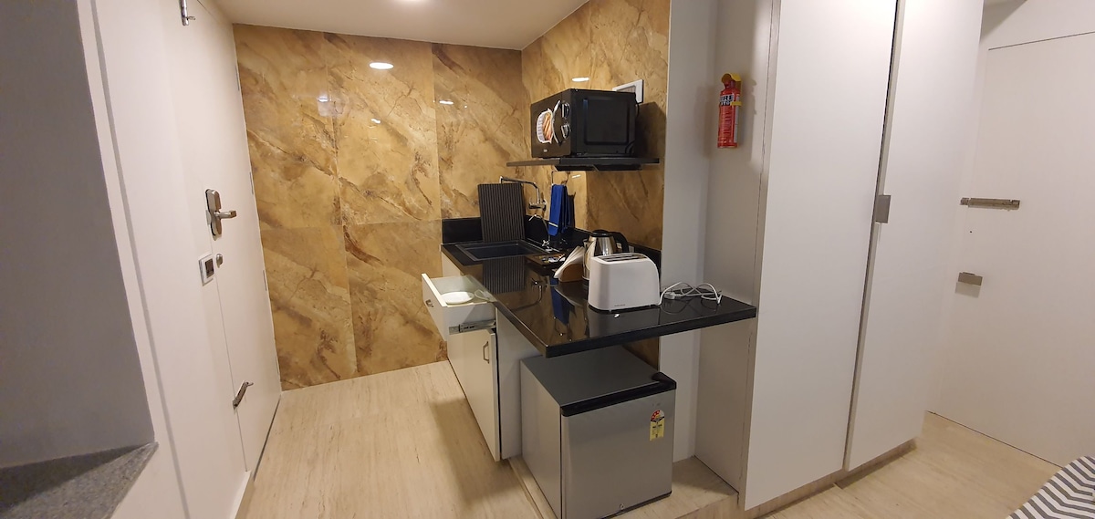 A modern kitchenette is shown, featuring a sleek black countertop with a microwave, toaster, and mini-fridge. The backsplash has a warm, marbled design, complementing the light-toned flooring. Cabinets provide storage, while ample lighting enhances the workspace's functionality.