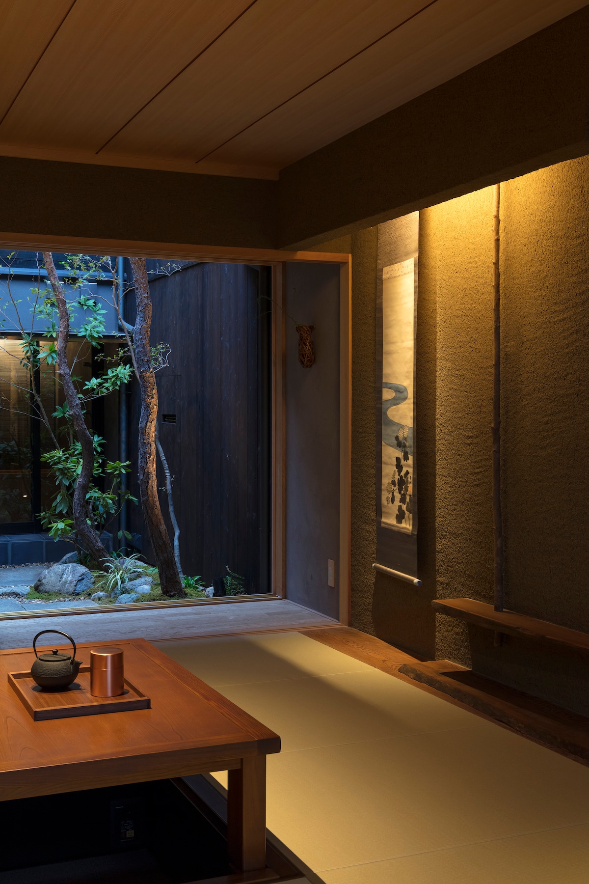 A serene Japanese-style room is illuminated by soft lighting, featuring a low wooden table with a kettle and cup. A view of the garden through the sliding door is framed by natural elements. Wooden planks and tatami flooring create a calming atmosphere.