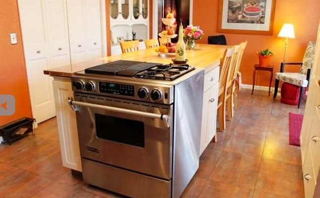 A modern kitchen features a stainless steel range with a five-burner cooktop set into an island with a wooden countertop. Light oak cabinets provide ample storage, while a dining area is defined by several wooden chairs around the table. Warm orange walls create a welcoming environment.