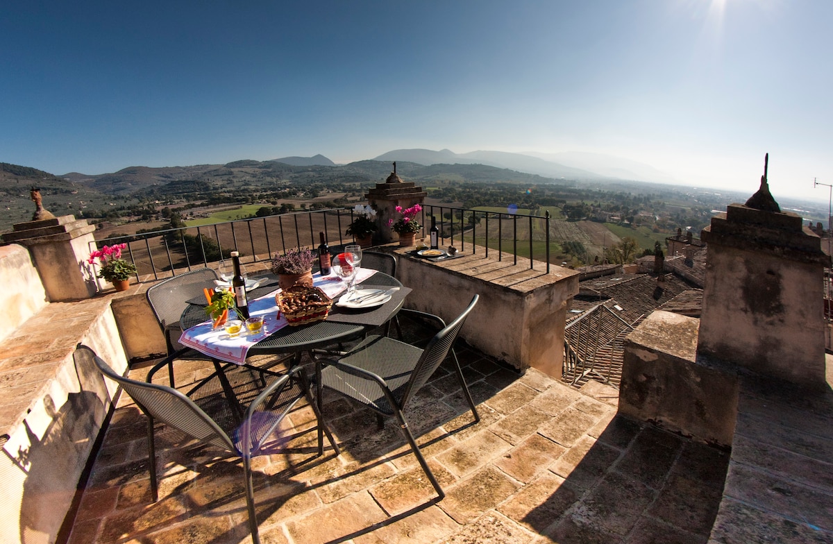 A terrace atop a historic building offers an outdoor dining space with a round table and four chairs. A stunning view of the Umbrian countryside extends beyond, with rolling hills and distant mountains visible under a clear blue sky.
