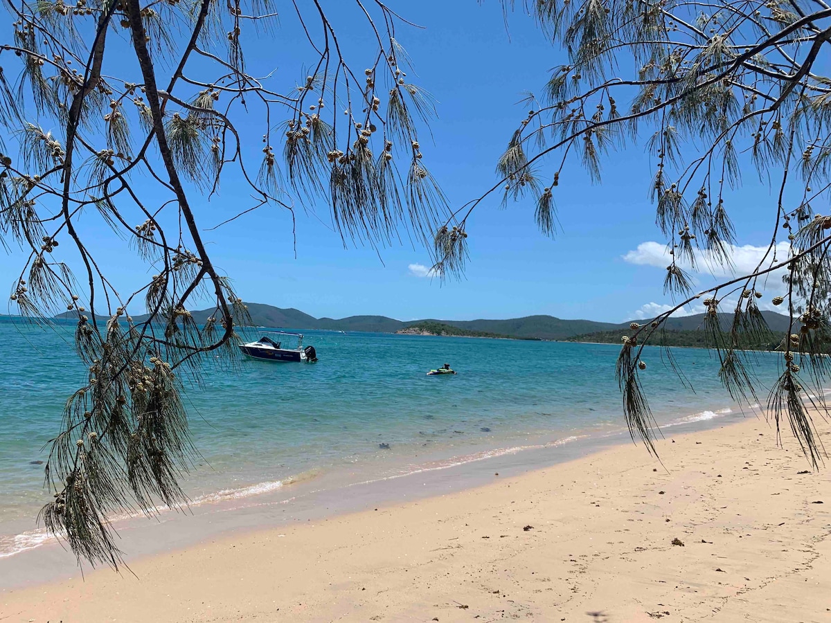 A serene beach scene is captured through branches of a tree, revealing soft sand and clear turquoise waters. A small boat is anchored in the distance, while gentle waves lap at the shore, showcasing the peaceful atmosphere of Hideaway Bay.