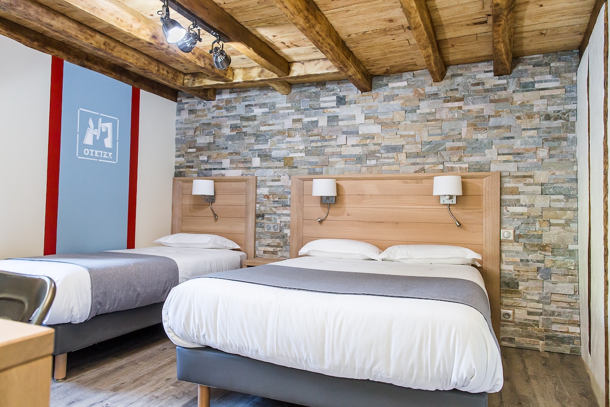 A cozy room features two beds: a spacious double bed and a comfortable single bed, both with light bedding. The wood-beamed ceiling adds character, while a stone accent wall provides a rustic touch. Soft lighting enhances the warm ambiance of the space.