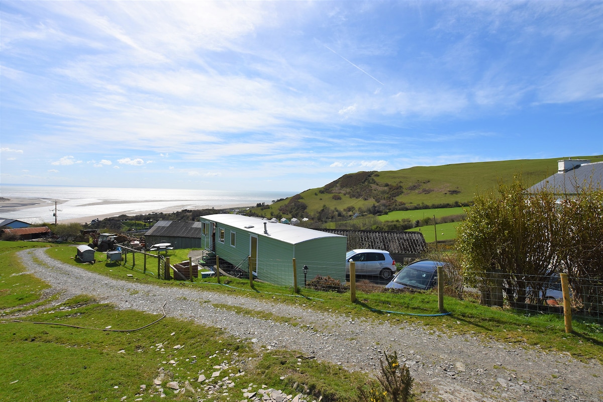 A scenic view captures the caravan set along a gravel path, surrounded by lush green fields. The coastline stretches in the background, meeting the sandy shore and sea under a clear blue sky. Nearby vehicles and a gentle rise in the land enhance the tranquil rural setting.