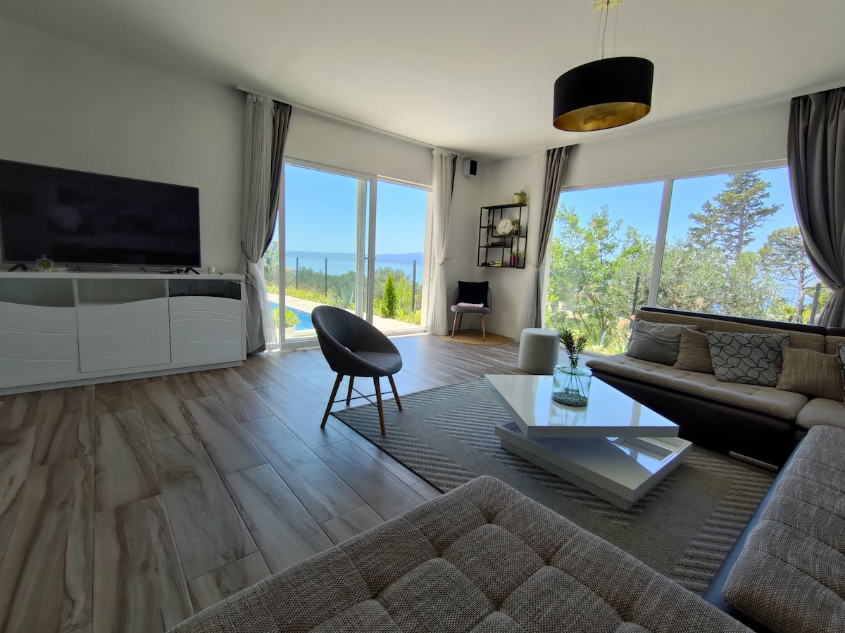 A contemporary living area features large windows that provide natural light and views of the surrounding landscape. A spacious sectional sofa is complemented by a sleek coffee table, with a minimalist television unit positioned against a wall. A small desk is placed beside the windows.