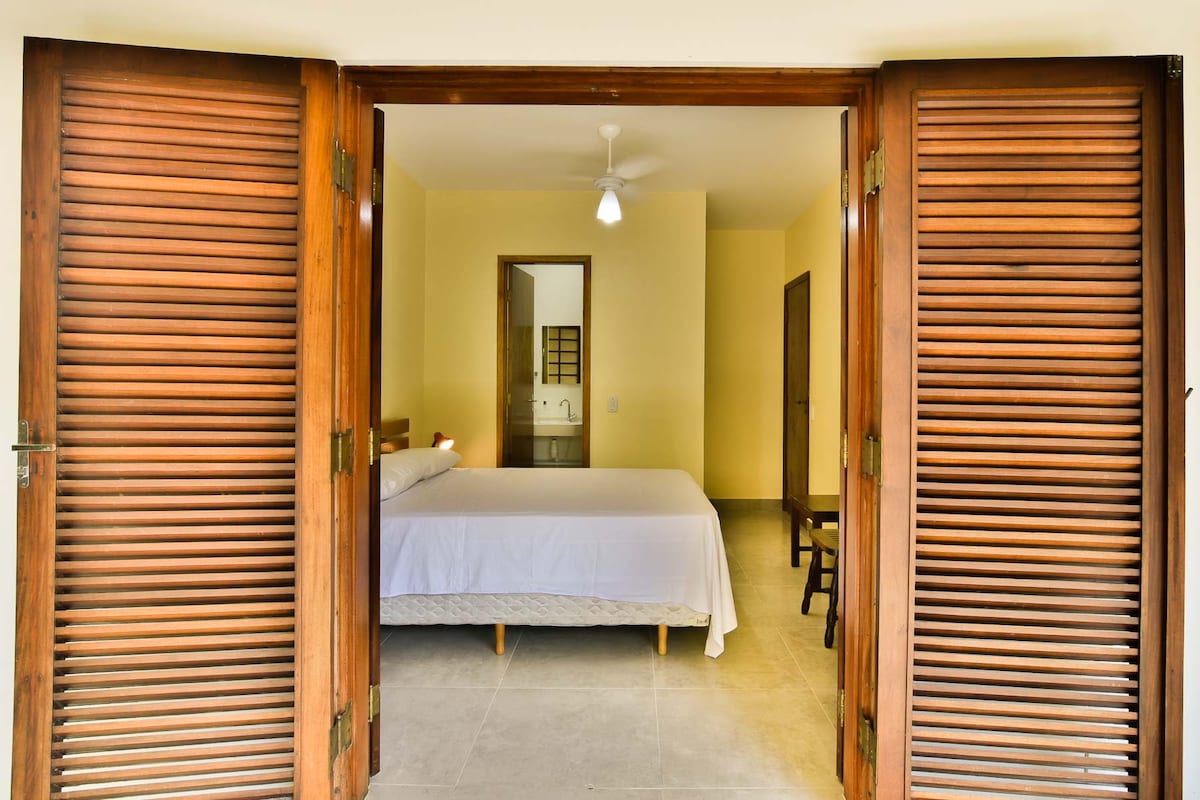 A spacious room is entered through wooden double doors, which are partially open. A large bed with a white coverlet occupies the center of the room, along with a ceiling fan above. A simple wooden chair and a doorway leading to a bathroom are also visible.