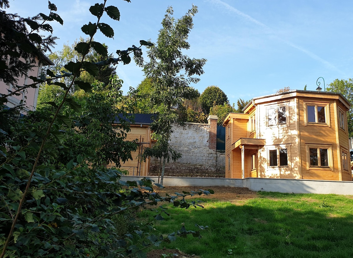 A wooden chalet, built in 2019, is situated within a private park. The structure features large windows and a porch, surrounded by greenery. Nearby, a neighboring wall is visible, providing a sense of privacy in the natural setting.