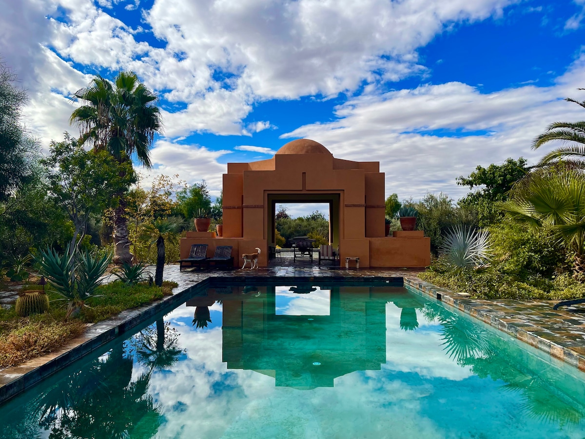 A serene swimming pool reflects the clear blue sky and surrounding greenery. An architectural feature with a curved roof stands in the background, framed by lush plants and palm trees. A seating area is visible, offering a peaceful space for relaxation.