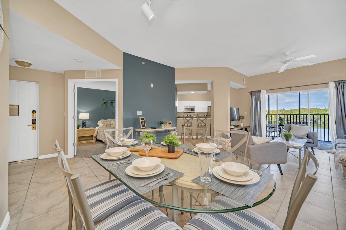 A dining area features a glass table set with plates and glasses, surrounded by striped chairs. The open layout connects to a cozy living space, visible in the background, with a view of the lake through sliding glass doors.