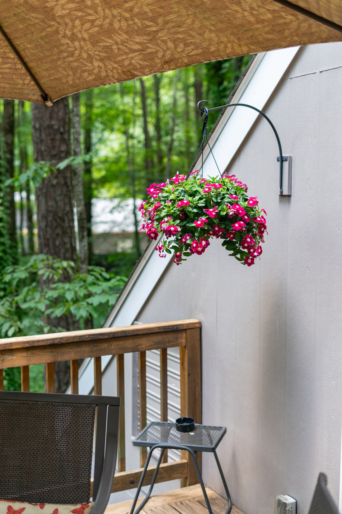 A hanging floral basket filled with vibrant pink flowers is displayed on a patio, complemented by a wooden railing and a small table. A shaded area is provided by a large umbrella, with greenery from trees visible in the background.