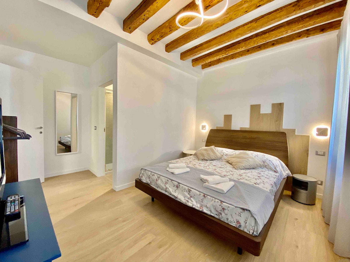 A calm bedroom features a spacious bed with a floral-patterned duvet. Soft lighting is provided by wall sconces, alongside wooden beams on the ceiling. A mirrored wardrobe is visible, while a door opens to the adjoining bathroom. Natural light filters through a sheer window covering.