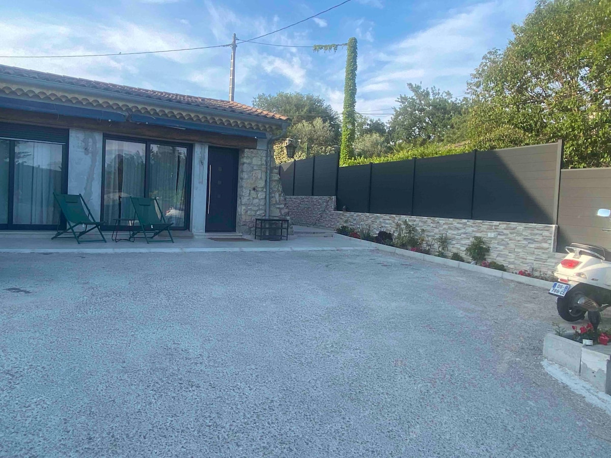 A spacious outdoor area is visible, featuring a stone exterior and a level parking space suitable for multiple vehicles. Two green chairs are positioned on the patio, alongside a small table. Lush greenery and a low stone wall surround the property, enhancing the serene ambiance.