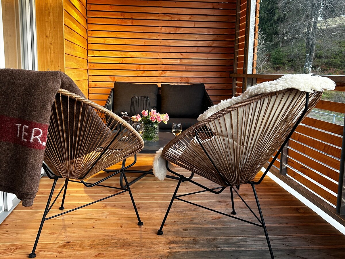 A balcony area is featured with two stylish chairs positioned around a low table. A cozy throw blanket drapes one chair, while a pot of flowers and two glasses rest on the table. Natural wood panels create a warm atmosphere.