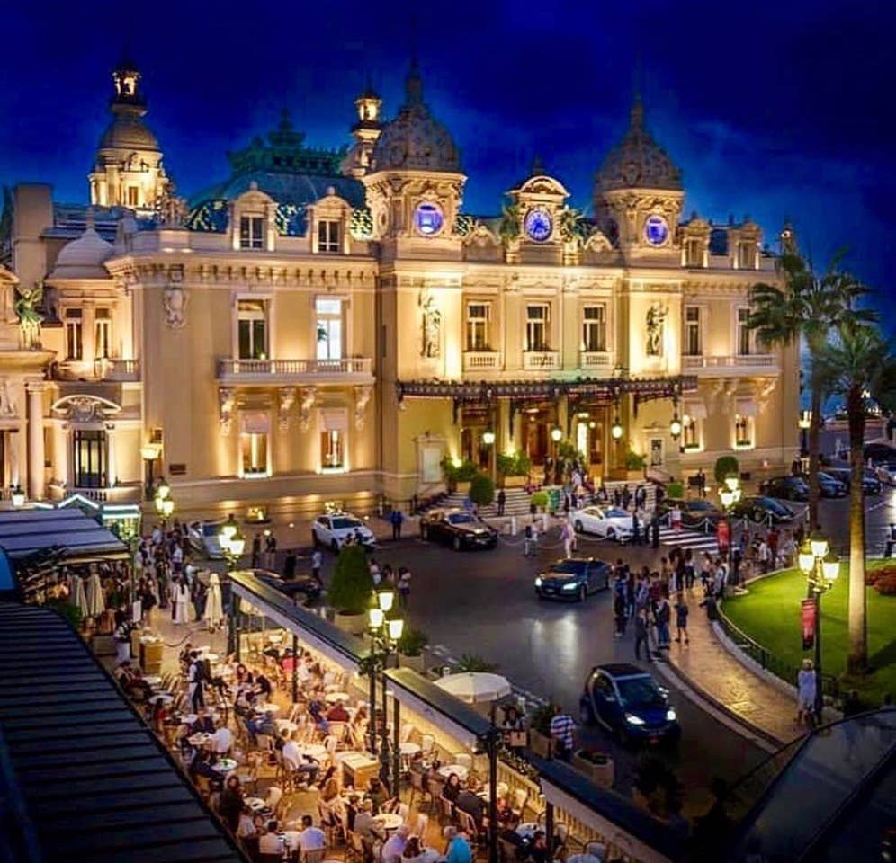 The iconic Casino de Monte-Carlo is illuminated at night, showcasing its ornate architectural features. A bustling atmosphere is visible with guests dining outdoors and cars parked in front. Lush palm trees accentuate the tropical environment, enhancing the elegance of this renowned landmark.