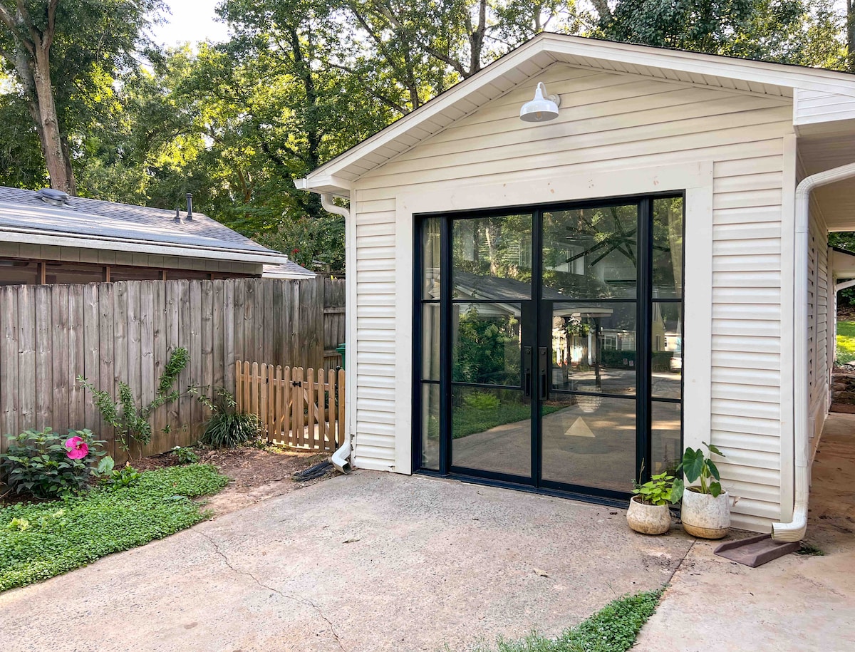 The exterior of a guest house is shown, featuring large glass doors that provide a view of the surrounding greenery. A small garden area is visible, with a flowering plant and potted greenery positioned near the entrance. The driveway is paved, offering space for parking.