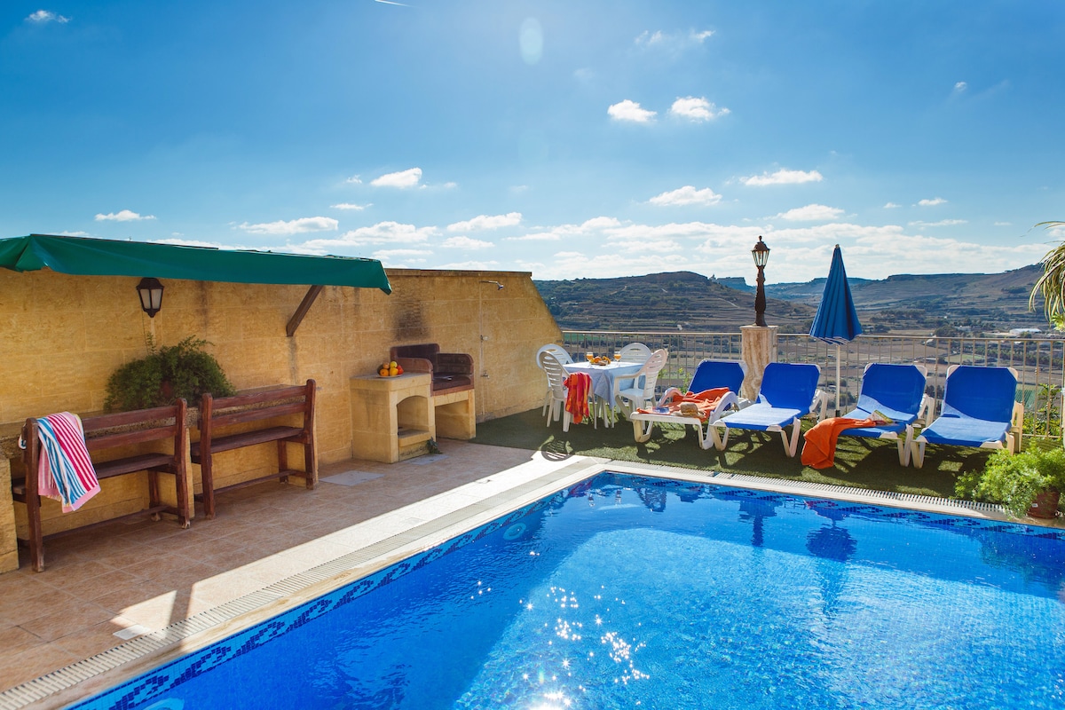A private pool area is visible, featuring a clear blue pool surrounded by sun loungers and a shaded seating area. Colorful towels are draped over the loungers, and a patio umbrella provides shade. Scenic views of the hills and sky are seen in the background.